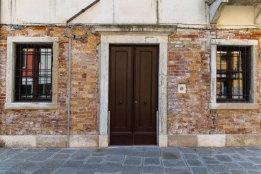 Parmaklıklı pencereleri ve karanlık kapısı olan eski tuğla bina. Sessiz bir şehir sokağında iki parmaklıklı pencereyle çevrili koyu ahşap kapılı tarihi bir tuğla cephesinin simetrik görüntüsü