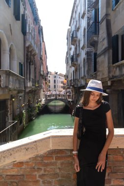 Venice Kanal Köprüsü 'nde Siyah Elbiseli Genç Kadın. Siyah elbiseli ve şapkalı şık bir genç kadın Venedik köprüsünde duruyor, tarihi binalar ve arka planda kalabalık bir yaya köprüsü olan bir kanala bakıyor..