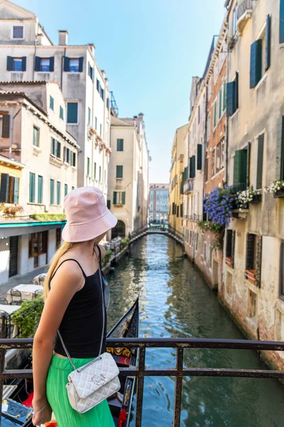 Turist kadın Venedik kanal manzarasını seyrediyor. Genç bayan gezgin, gondolları ve renkli tarihi binaları olan dar bir Venedik kanalına bakan bir köprüde duruyor.
