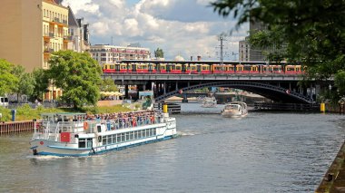 Berlin, Almanya, 26 Mayıs 2025: Berlin Şehri 'nde Tur Tekneleri ve Tren Geçidi Köprüsü. Güneşli bir günde Berlin 'in merkezindeki bir köprüden sarı-kırmızı bir tren geçerken Spree Nehri' nde gezinti yapan tekneleri görmek..