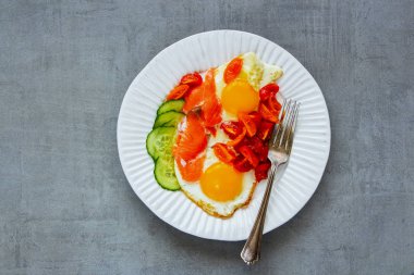 Sabah kahvaltı plaka kapatın. Som balığı, kızarmış yumurta, domates ve salatalık açık beton renkli üstten görünüm üzerinde füme. İyi yağlar sağlıklı beslenme kavramı. Düz yatıyordu stili.