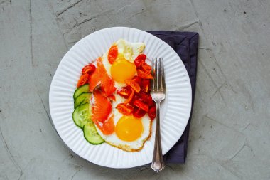 Morning breakfast in white plate flat-lay. Smoked salmon, fried eggs, tomatoes and cucumber over light concrete background top view. Good fats healthy eating concept. clipart