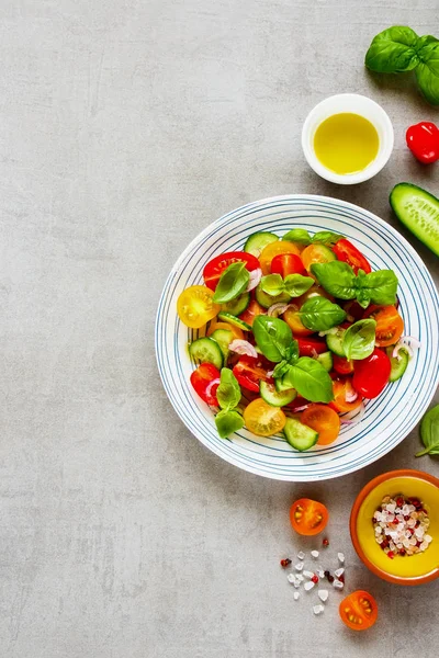 Healthy salad and ingredients on light background from above. Fresh cherry colorful tomatoes, cucumbers, red onion, olive oil, basil and spices flat lay.