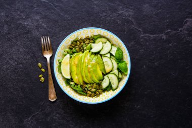 Yeşil vegan salata kasesi avokado, roka, salatalık ve kabak çekirdeği siyah arka plan üzerine kapatın. Üstten görünüm, düz yatıyordu stili.