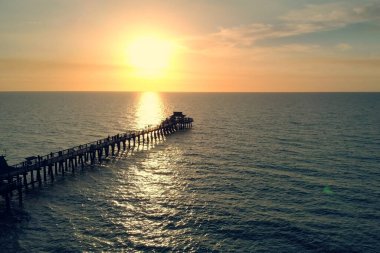Silhouette of a pier over the water at sunset. Naples Beach and Fishing Pier at Sunset, Florida. Small Pier in Calm Ocean with Sun Reflecting on Water. Sunset over the Pier. Beautiful Ocean Sunsets. clipart