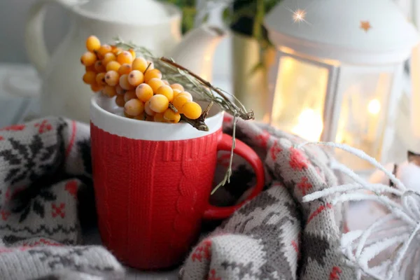WINTER COZY BACKGROUND - RED CUP OF TEA WITH ORNAMENT, WOOL SCARF, FLASHLIGHT, KETTLE, BRANCH OF YELLOW BERRIES