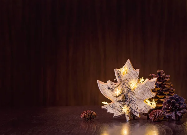 Simple, natural Christmas decor of pine cones with rustic maple leaf ornament wrapped in lights on a dark wood background.