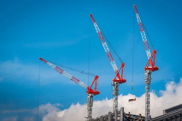 Three construction tower cranes operation at a construction site against blue sky background.