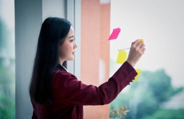 Genç Asyalı iş kadınının portresi modern ofiste cama yapışkan kağıt bloknot yazıyor. İş planı konsepti.