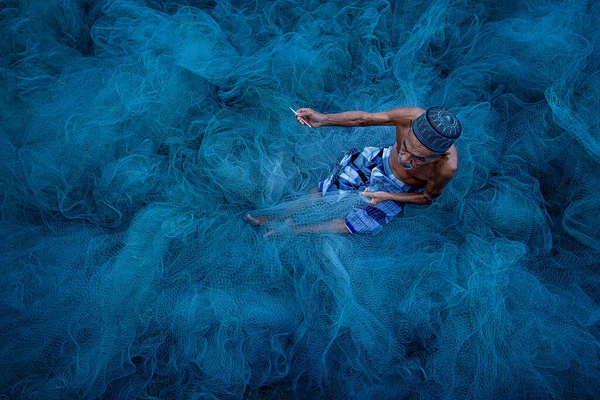 Nakorn Si Thammarat, Thailand - August 17, 2020 : Old fisherman hands sewing blue fishing nets sitting on the ground and surrounded big net — Stock Image Nakorn Thammarat Thailand August 2020 Old Fisherman Hands Sewing Blue Stock Photo