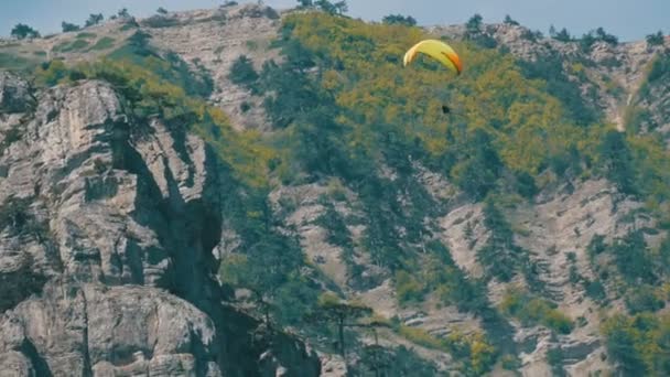 Gelber Gleitschirm mit orangefarbenen Streifen fliegt in einer wunderschönen Bergregion vor dem Hintergrund grauer großer Felsen — Stockvideo