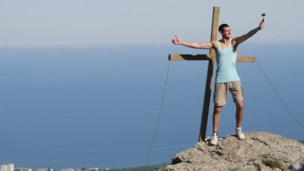 Giovane uomo si toglie con gioia sulla macchina fotografica d'azione, in piedi sulla cima della montagna vicino alla croce. Raggiungere l'obiettivo, la motivazione — Video Stock