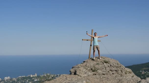 Mladý muž radostně bere sám na akční kamery, stojící na vrcholu hory v blízkosti kříže. Dosažení cíle, motivace — Stock video