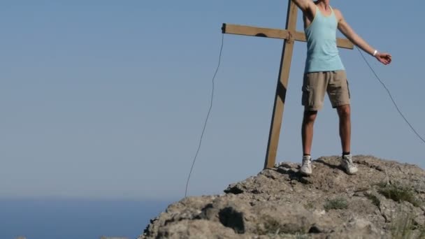 Le jeune homme s'enlève joyeusement sur la caméra d'action, debout au sommet de la montagne près de la croix. Atteindre l'objectif, la motivation — Video