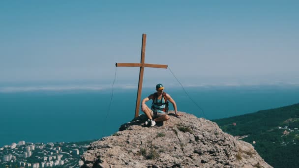 Sırtında bir sırt çantası ile gezgin adam dağın zirvesinde bir haç ile tepesinde oturuyor. Başarı hedefi, motivasyon yükselmeye. — Stok video