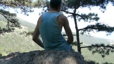 Genç bir adam bir yükseltilmiş platform üzerinde oturur ve güzel dağ manzarası mesafe, görünüm geri gelen görünüyor