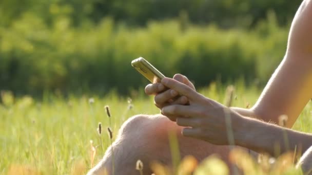 Joven mira el teléfono inteligente y algo se imprime en la hierba verde sobre el telón de fondo de una hermosa naturaleza de verano — Vídeos de Stock