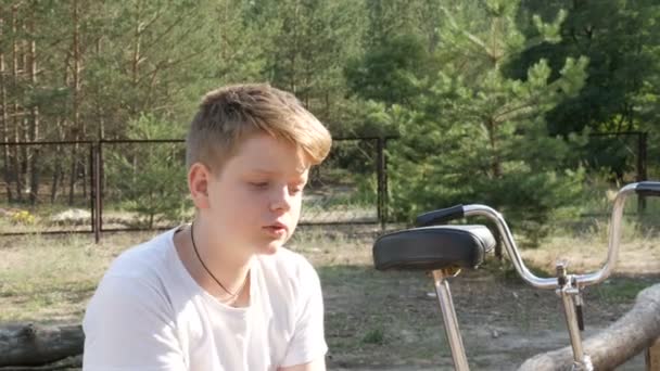 Blondă drăguță cu ochi albaștri un băiat adolescent stă într-un parc lângă bicicletă și vorbește vara. Portretul emoțional al unui băiat — Videoclip de stoc