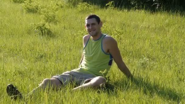 Retrato de un joven guapo y alegre hombre sentado en la hierba en la naturaleza en verano sobre el fondo del bosque y mirando a su alrededor — Vídeos de Stock