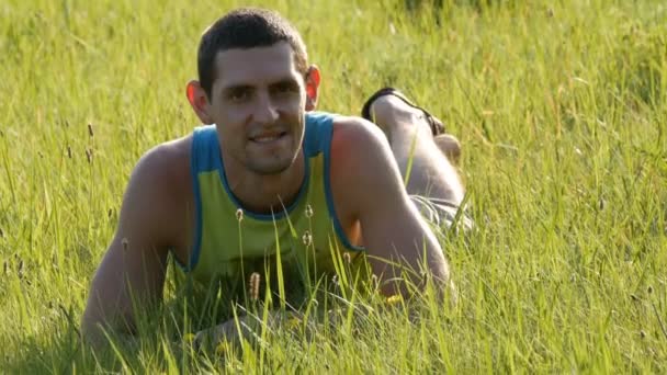 Vrolijke jongeman zit op een groen gras in de zomer en glimlacht — Stockvideo