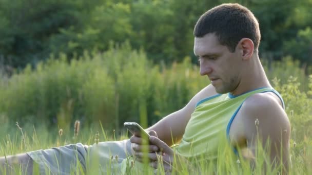Молодий чоловік набирає щось на свій смартфон, сидячи на природі на зеленій траві влітку — стокове відео