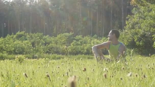 Веселый молодой человек летом сидит на зеленой траве и улыбается. — стоковое видео