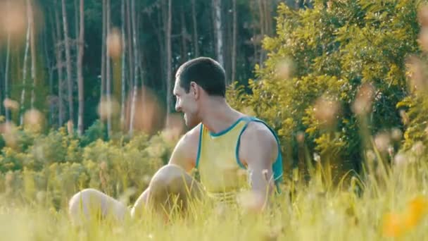 Χαρούμενος νεαρός άνδρας κάθεται σε ένα πράσινο γρασίδι το καλοκαίρι και χαμόγελα — Αρχείο Βίντεο