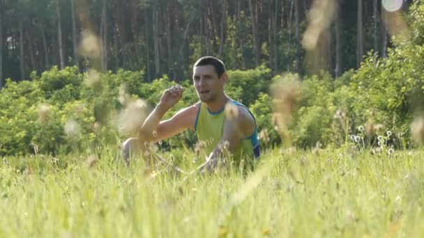 Портрет молодого красивого веселого человека, сидящего летом на траве на фоне леса и оглядывающегося вокруг — стоковое видео