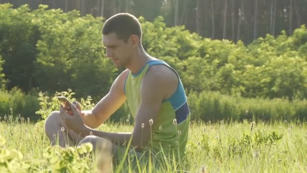 Jovem está digitando algo em seu smartphone enquanto sentado na natureza na grama verde no verão — Vídeo de Stock