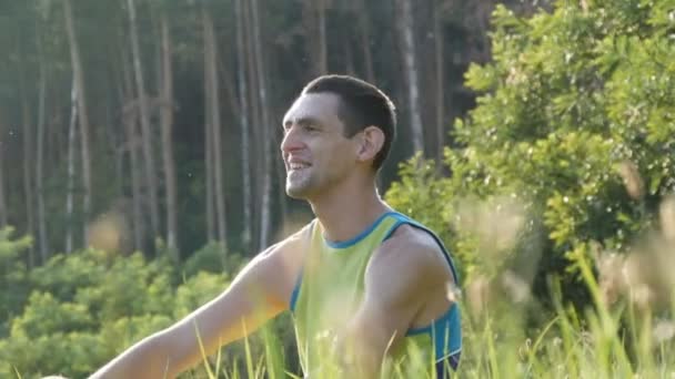 Молодой человек сидит на природе в траве счастливо наслаждаясь жизнью и улыбаясь — стоковое видео
