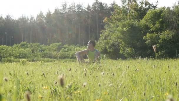 Портрет молодого красивого веселого человека, сидящего летом на траве на фоне леса и оглядывающегося вокруг — стоковое видео