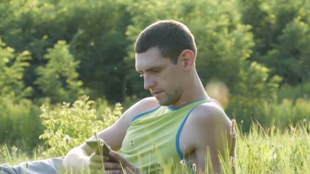 Молодий чоловік набирає щось на свій смартфон, сидячи на природі на зеленій траві влітку — стокове відео