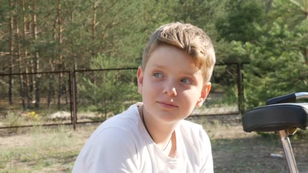 Мальчик-подросток сидит в лесном летнем парке рядом с велосипедом и смотрит в камеру — стоковое видео