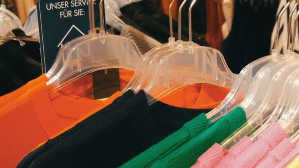 Various stylish knitted multicolored sweaters hanging on fashion black hangers in a clothing store in mall or shopping center. On the plate for the service inscription in German — Stock Video