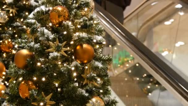 Beautifully decorated Christmas tree with large gold and silver balls, stars, garlands and artificial snow is standing in the shopping center or mall, people are passing by on the escalator — Stock Video