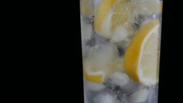 Limón en rodajas en un vaso largo con cubitos de hielo y refrescos fríos sobre un fondo negro vista de cerca. Refrescante Limonada Burbujas . — Vídeo de stock