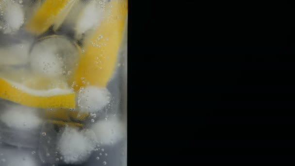 Nakrájené citrony v dlouhém skle s kostkami a studenou sodou na černém pozadí zblízka. Osvěžující bubliny na limonádě. — Stock video