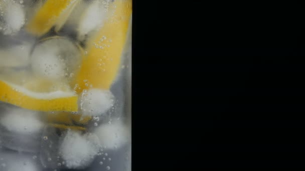 Limón en rodajas en un vaso largo con cubitos de hielo y refrescos fríos sobre un fondo negro vista de cerca. Refrescante Limonada Burbujas . — Vídeo de stock