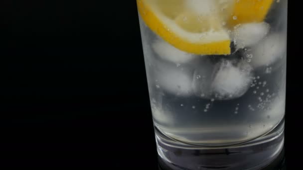 Gesneden citroen in een lang glas met ijsblokjes en koude soda op een zwarte achtergrond close-up te bekijken. Verfrissende limonade bubbels. — Stockvideo
