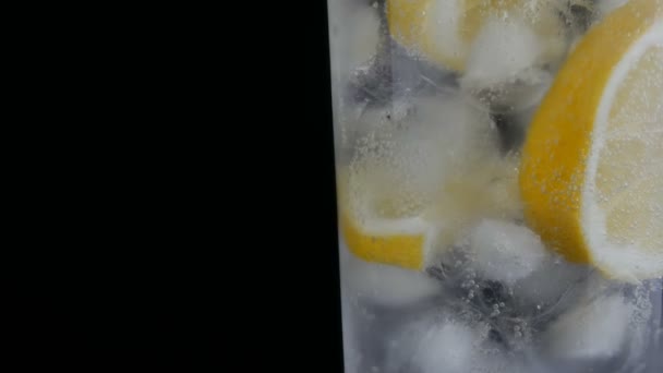 Sliced lemon in a long glass with ice cubes and cold soda on a black background close up view. Refreshing Lemonade Bubbles. — Stock Video