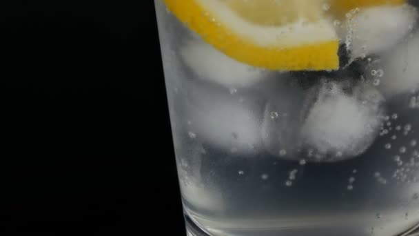 Limón en rodajas en un vaso largo con cubitos de hielo y refrescos fríos sobre un fondo negro vista de cerca. Refrescante Limonada Burbujas . — Vídeo de stock