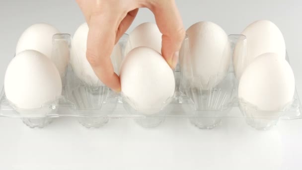 Nagy fehér csirke tojás átlátszó műanyag tálcát fehér alapon. Női kezek fel a tojásokat a tálcára — Stock videók