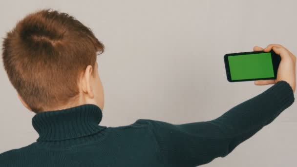 A teen boy stands with his back to the camera and is holding in hand a black smartphone with a green screen on white background. Teen Selfie — Stock Video © Milkare #251245638