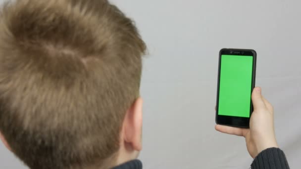 Ένα εφηβικό αγόρι στέκεται με την πλάτη του στην κάμερα και κρατά στο χέρι ένα μαύρο smartphone με μια πράσινη οθόνη σε λευκό φόντο. Έφηβος/η selfie — Αρχείο Βίντεο