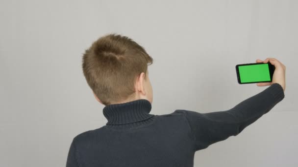 十代の少年は、カメラに彼の背中で立って、手に白の背景に緑の画面で黒のスマートフォンを保持しています。ティーンセルフ — ストック動画