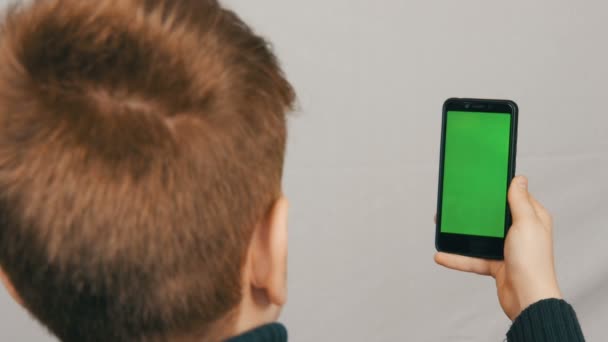 Ένα εφηβικό αγόρι στέκεται με την πλάτη του στην κάμερα και κρατά στο χέρι ένα μαύρο smartphone με μια πράσινη οθόνη σε λευκό φόντο. Έφηβος/η selfie — Αρχείο Βίντεο