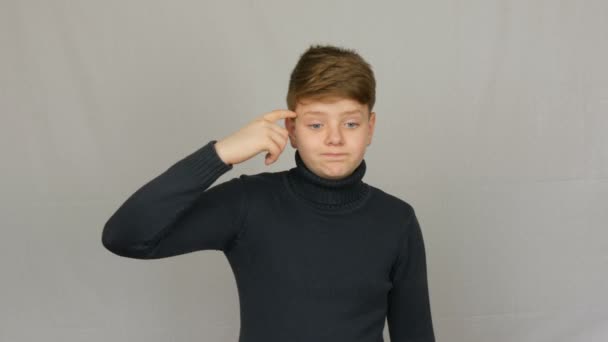 Πορτραίτο έφηβο αγόρι αναστρέφει το δάχτυλό του στο ναό του και φαίνεται κατηγοριμένο σε ένα λευκό φόντο στο στούντιο. Τα συναισθήματα των εφήβων — Αρχείο Βίντεο
