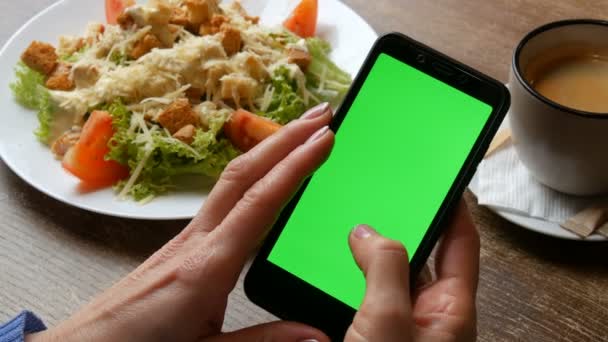 Tecla de croma o pantalla verde en un teléfono inteligente negro en manos femeninas con una manicura bien arreglada en el fondo de una ensalada César y una taza de café en un restaurante — Vídeo de stock