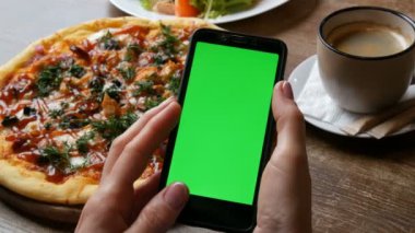 Büyük bir pizza ve kahve fincan arka planda iyi bakımlı manikür ile kadın elleri siyah bir smartphone üzerinde Chroma anahtar veya yeşil ekran
