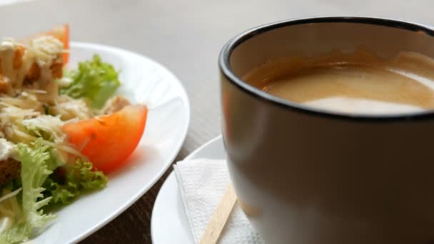 Вкусный свежий салат Цезарь с салатом, помидорами, кусками куриного мяса, сыром пармезан, крекерами и соусом на белой тарелке возле чашки кофе в кафе — стоковое видео